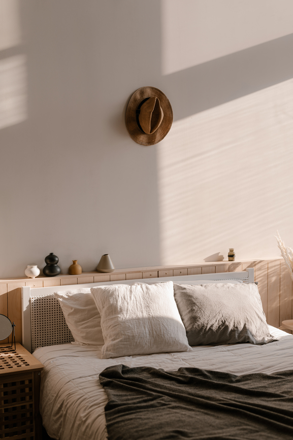 Close up shot in a bedroom of a wooden bed frame with white pillows, white sheets, and a grey comforter on top. A brown fedora hangs on the wall above the bed.