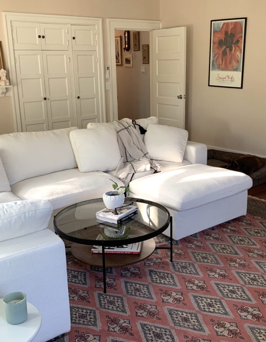 A living room with a white sectional sofa, a glass coffee table, a patterned rug, and a brown armchair. The walls are beige and decorated with framed artwork. A door and shelf are visible in the background.