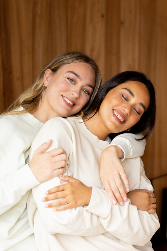 Two women embracing and smiling at the camera, one with blonde hair and the other with dark hair, both wearing white sweatshirts.