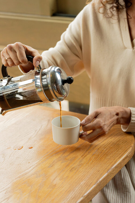 A person pours coffee from a french press into a white cup on a wooden table.