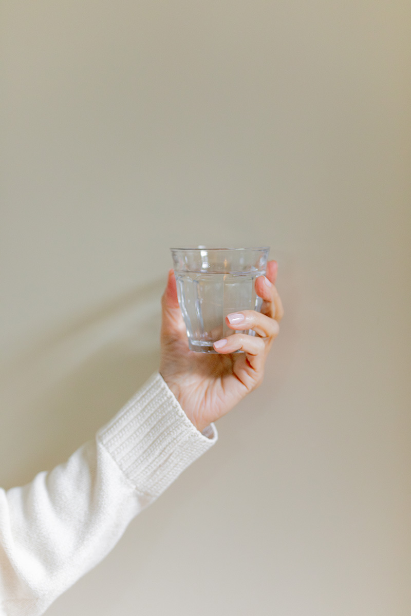 A person's hand holding a clear glass of water against a light beige background.