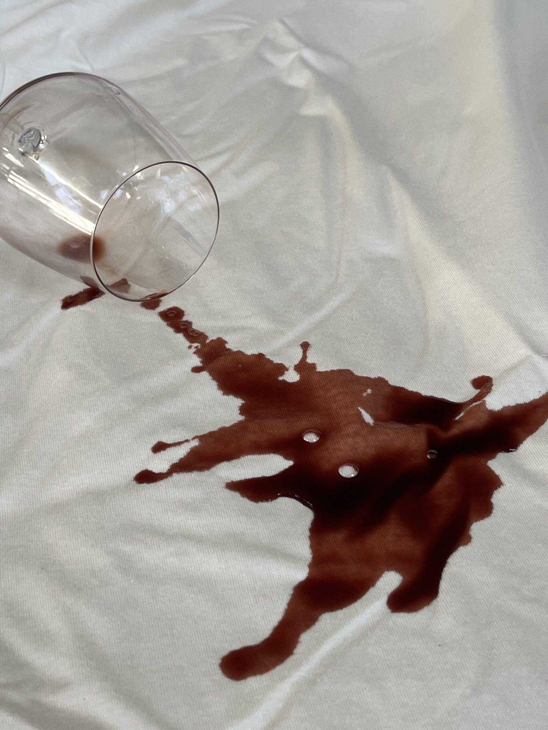 A wine glass lies on its side with spilled red wine leaking out onto a white mattress protector.