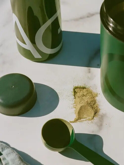 A close up of AG1 powder supplement, next to it's bottle and open container. 