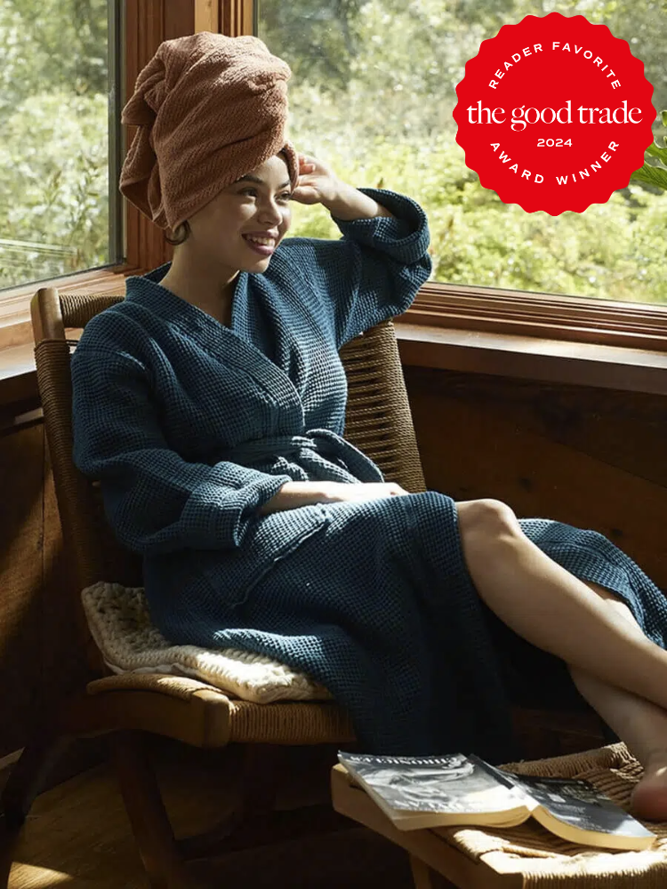 A model with her hair in a towel wearing a blue Coyuchi waffle knit robe while sitting on a wicker chair. The TGT 2024 Award Winner Badge is on the right corner of the image. 
