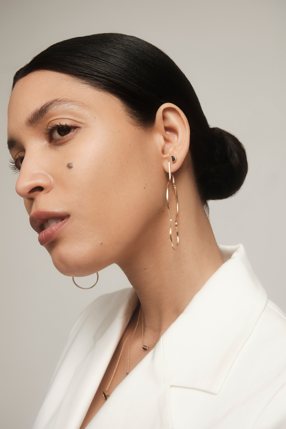A woman with sleek hair styled in a low bun, wearing hoop earrings and a white blouse, poses with her head turned to the side, showcasing her profile.