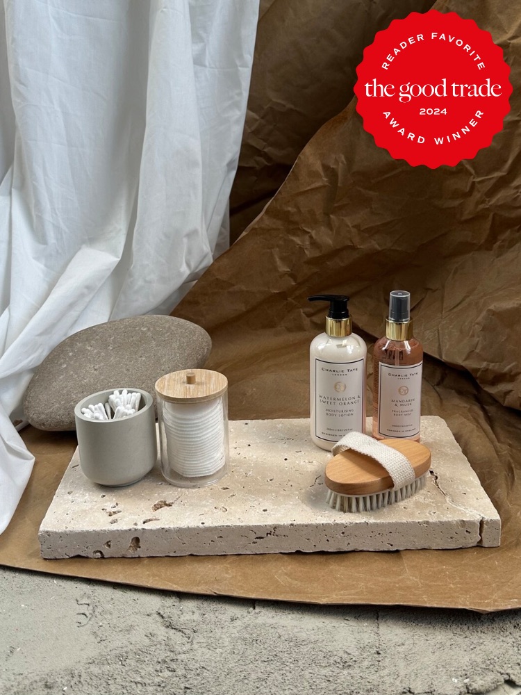 Two skincare product bottles with a brush and small containers on a concrete slab, against a draped fabric and stone background.