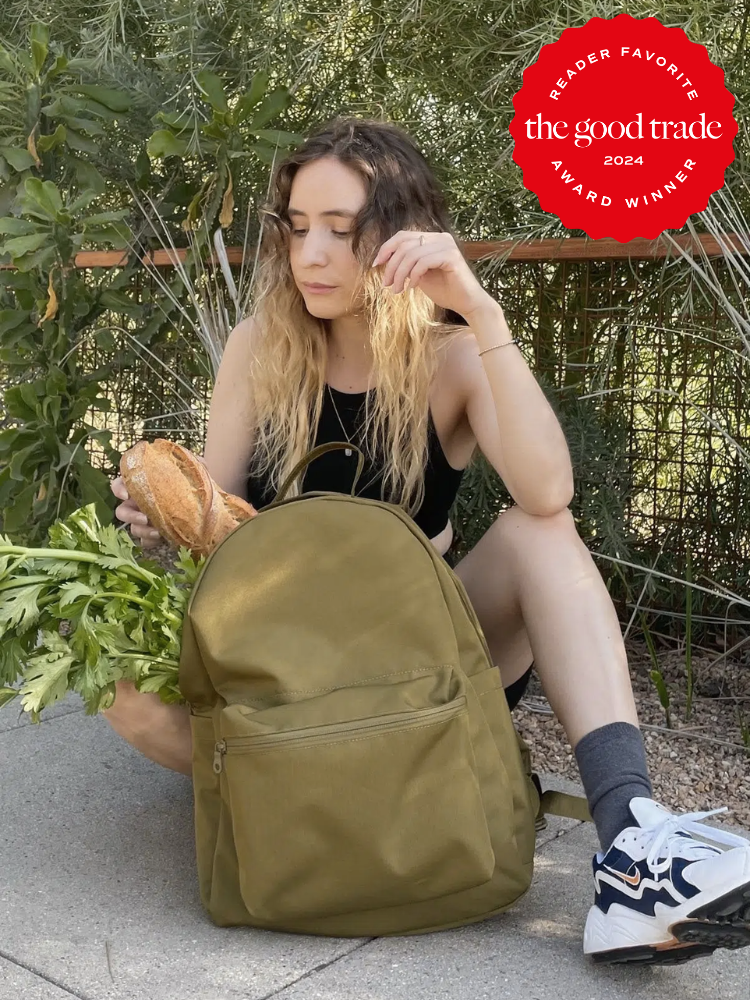 A model seated outside, removing a baguette from a Girlfriend Collective backpack. 
