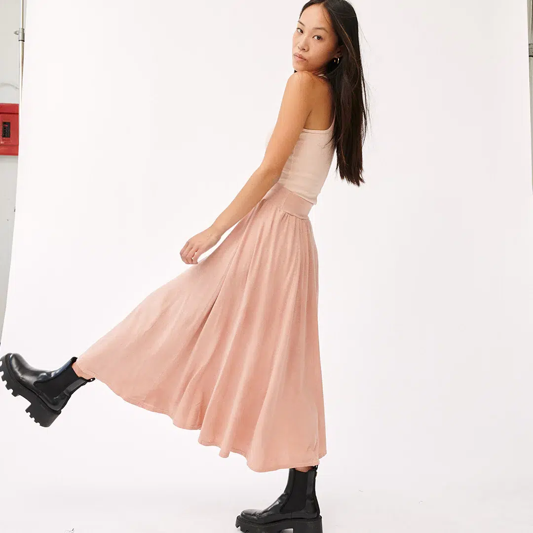 Young woman in a beige tank top and flowing skirt paired with black boots, playfully side-stepping against a white backdrop.