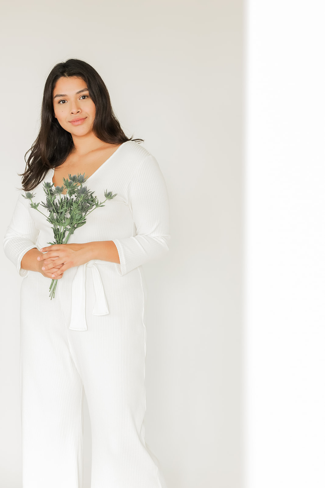 A woman in a white jumpsuit holds a bouquet of greenery, standing beside a white wall.