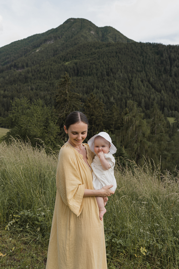 A mother holds a baby while standing in a mountainous, grassy field.