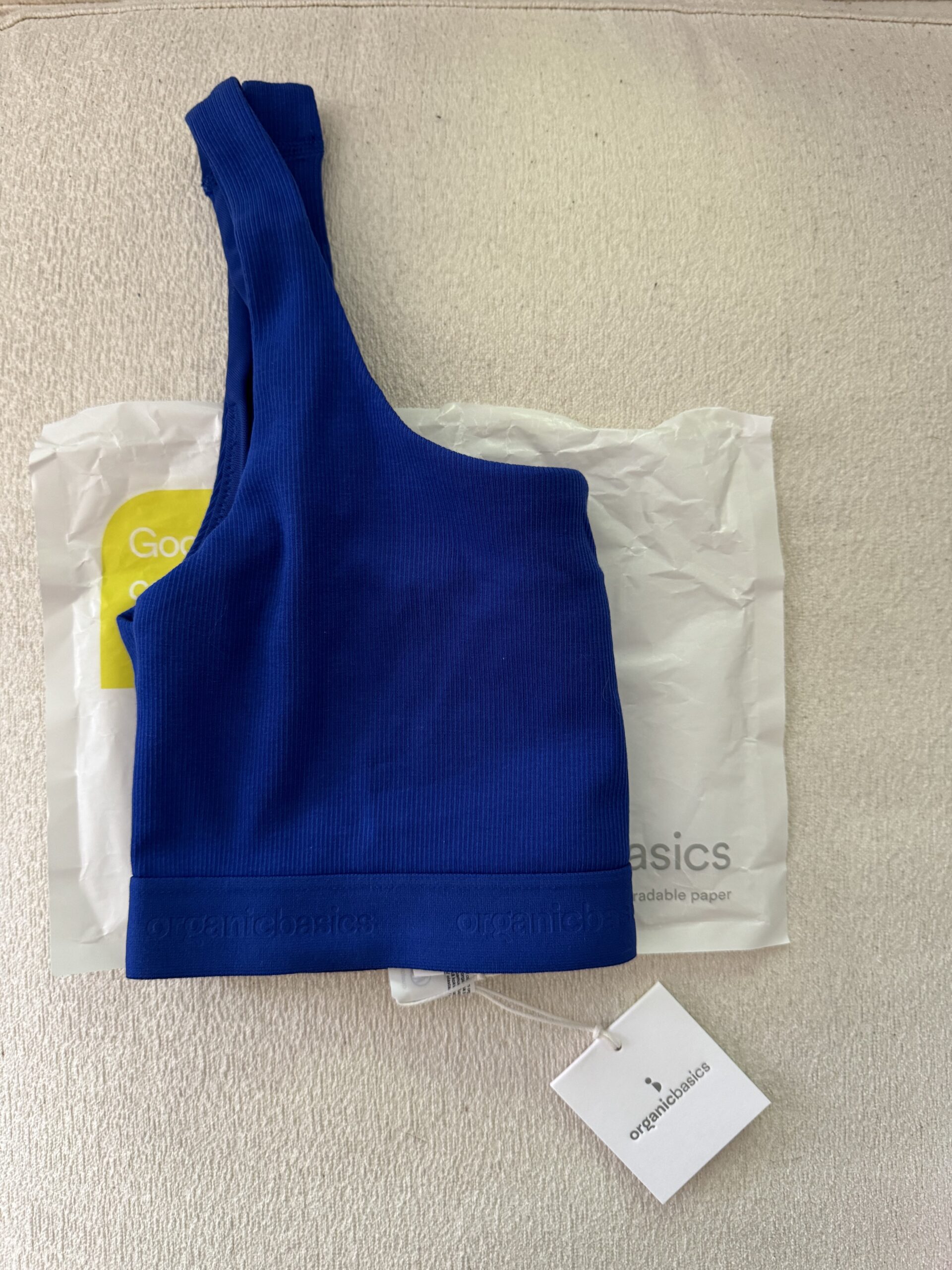 A blue one-shoulder top with a white tag laying on a white organic cotton diaper package, viewed from above.