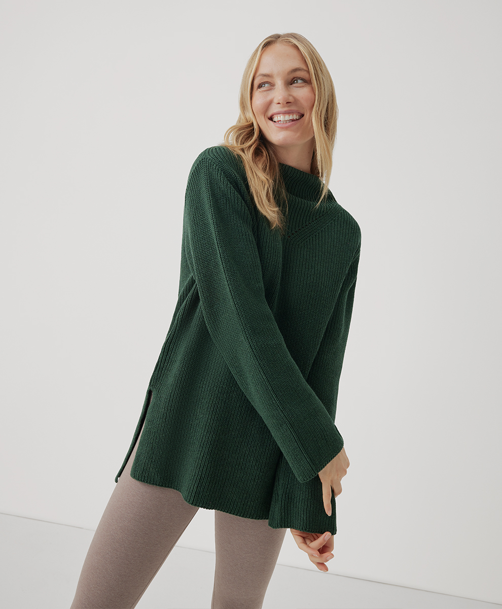 A model wearing a green Pact knit sweater and beige leggings.