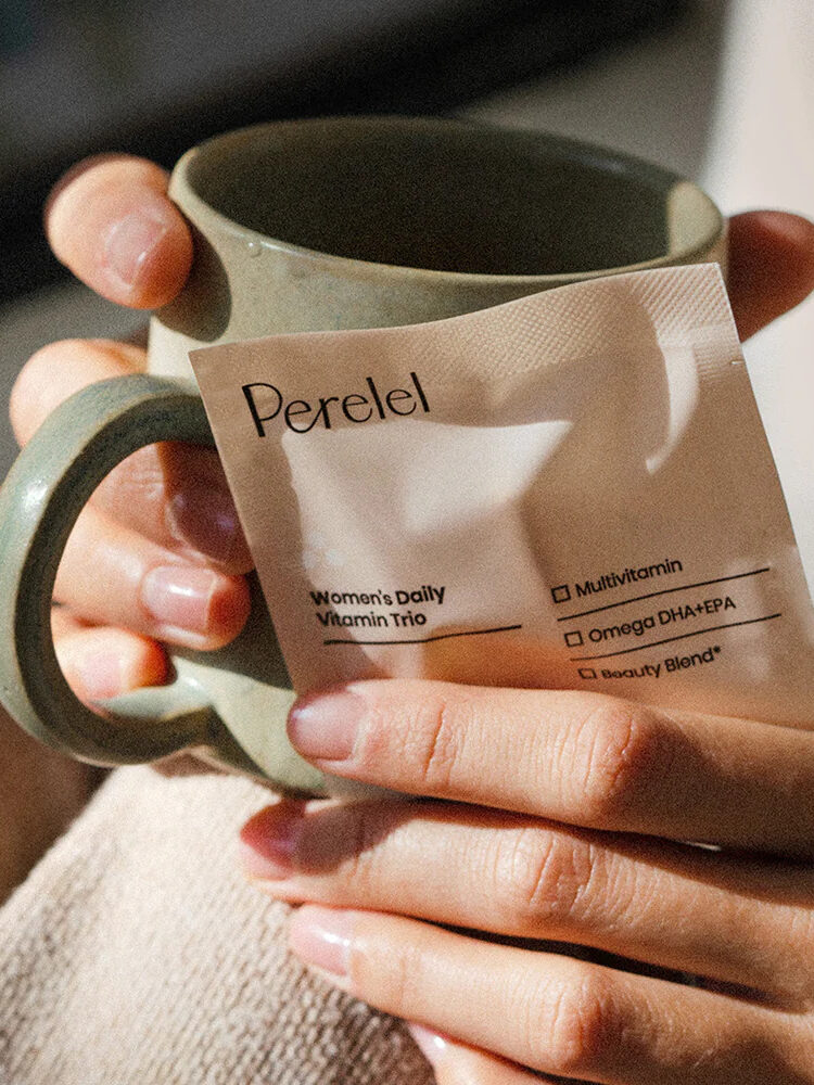 Hands holding a mug and a pack of Perelel Women's Daily Vitamin Trio.