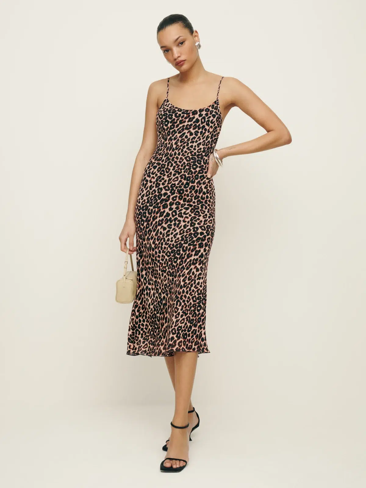 Woman posing in a leopard print dress with a small handbag and black heels.