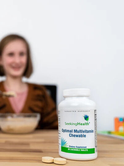 A bottle of Seeking Health Optimal Multivitamin Chewable tablets with a blurred model eating in the background.