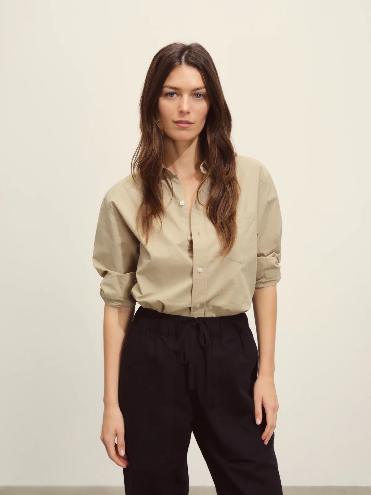 Woman in a beige blouse and black trousers standing against a neutral background.