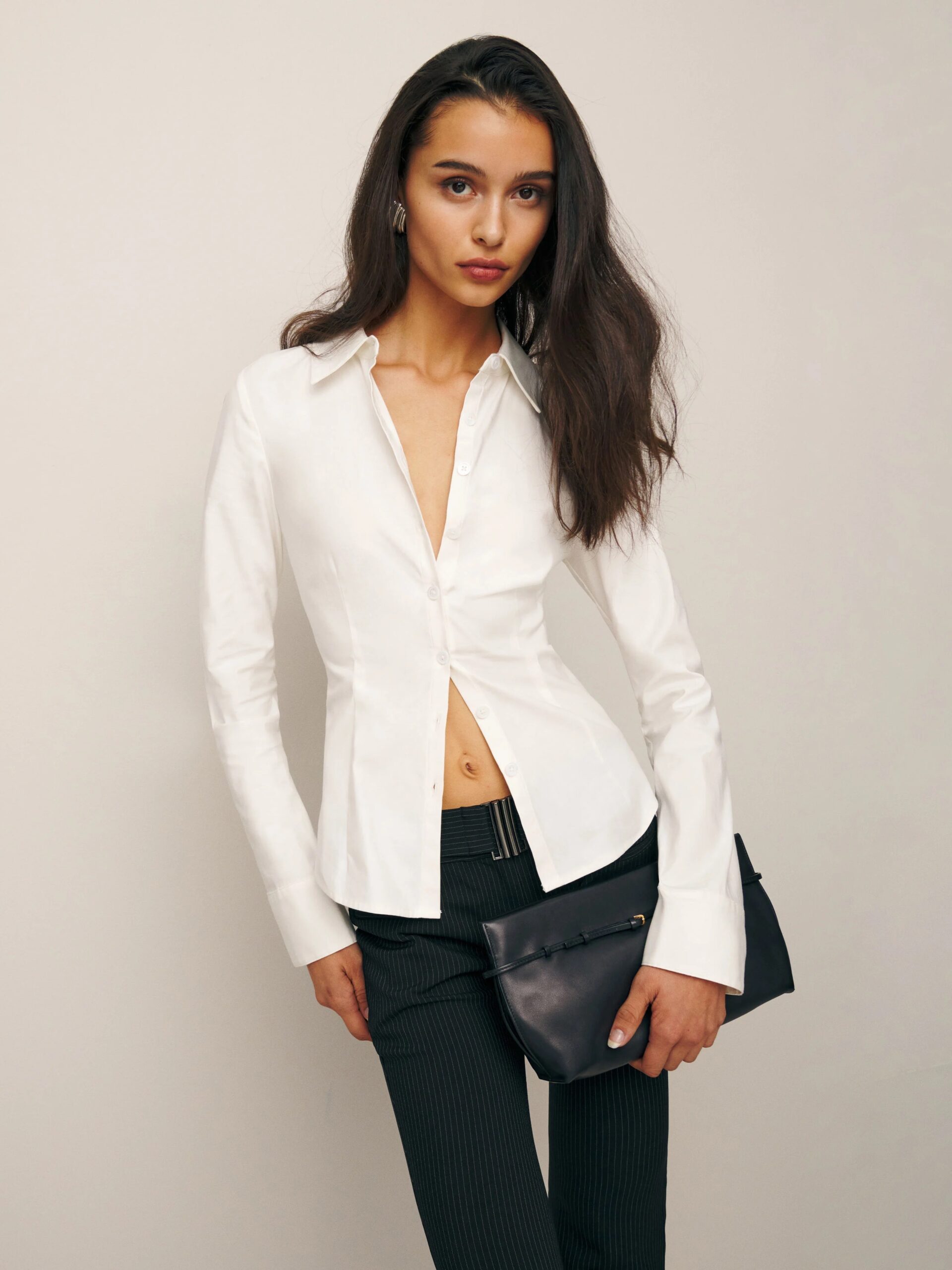 Woman posing in a white blouse and black trousers, holding a small bag.
