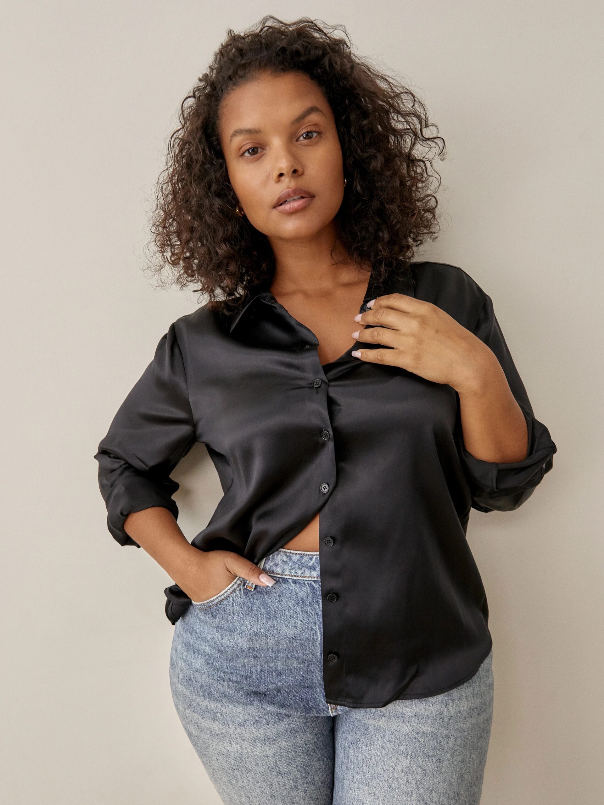 A woman with curly hair wearing a black shirt and blue jeans stands against a light backdrop, one hand partially tucked into her pocket.