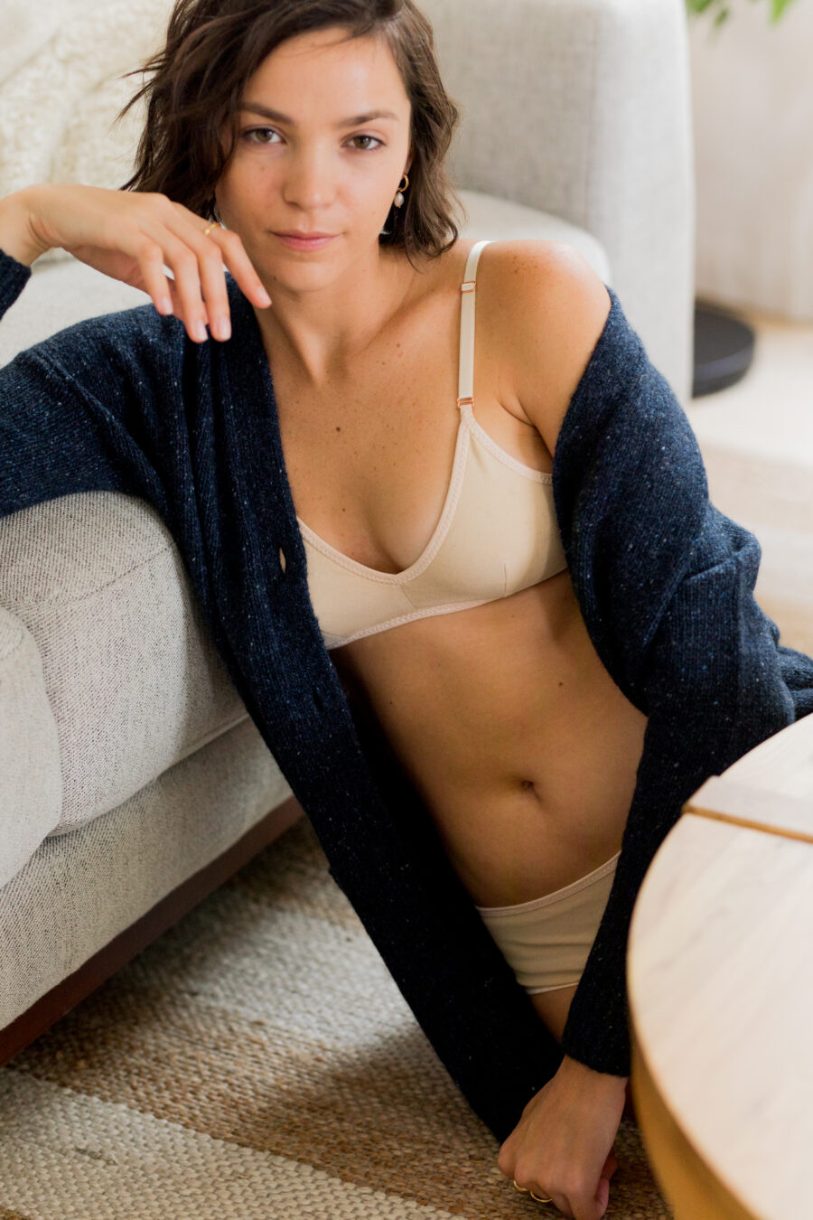 A model wearing organic cotton lingerie and a robe, while seated on he floor leaning against a couch.