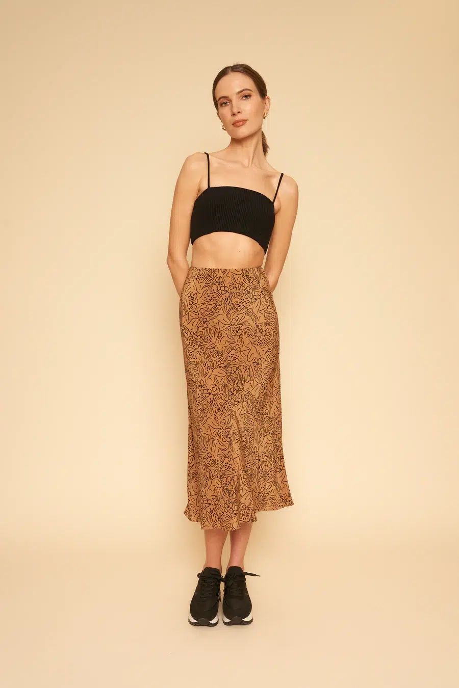 A woman in a black crop top and patterned skirt paired with black sneakers standing against a beige background.