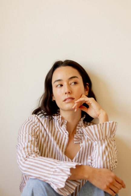 Woman with long dark hair sits against a plain light-colored wall, wearing a striped blouse and looking thoughtfully to the side with her hand resting near her face.