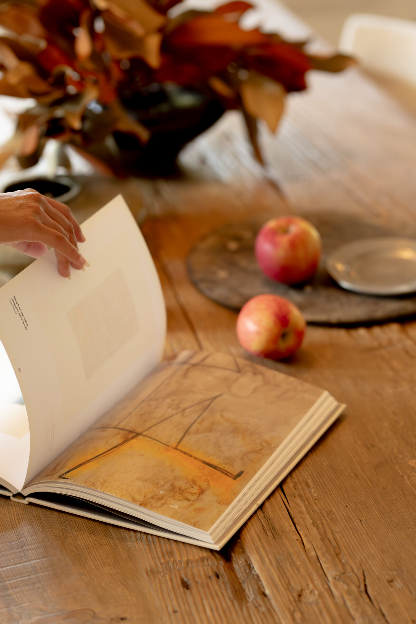 A person flips through an art book on a wooden table with two apples, a round tray, and dried leaves in the background.