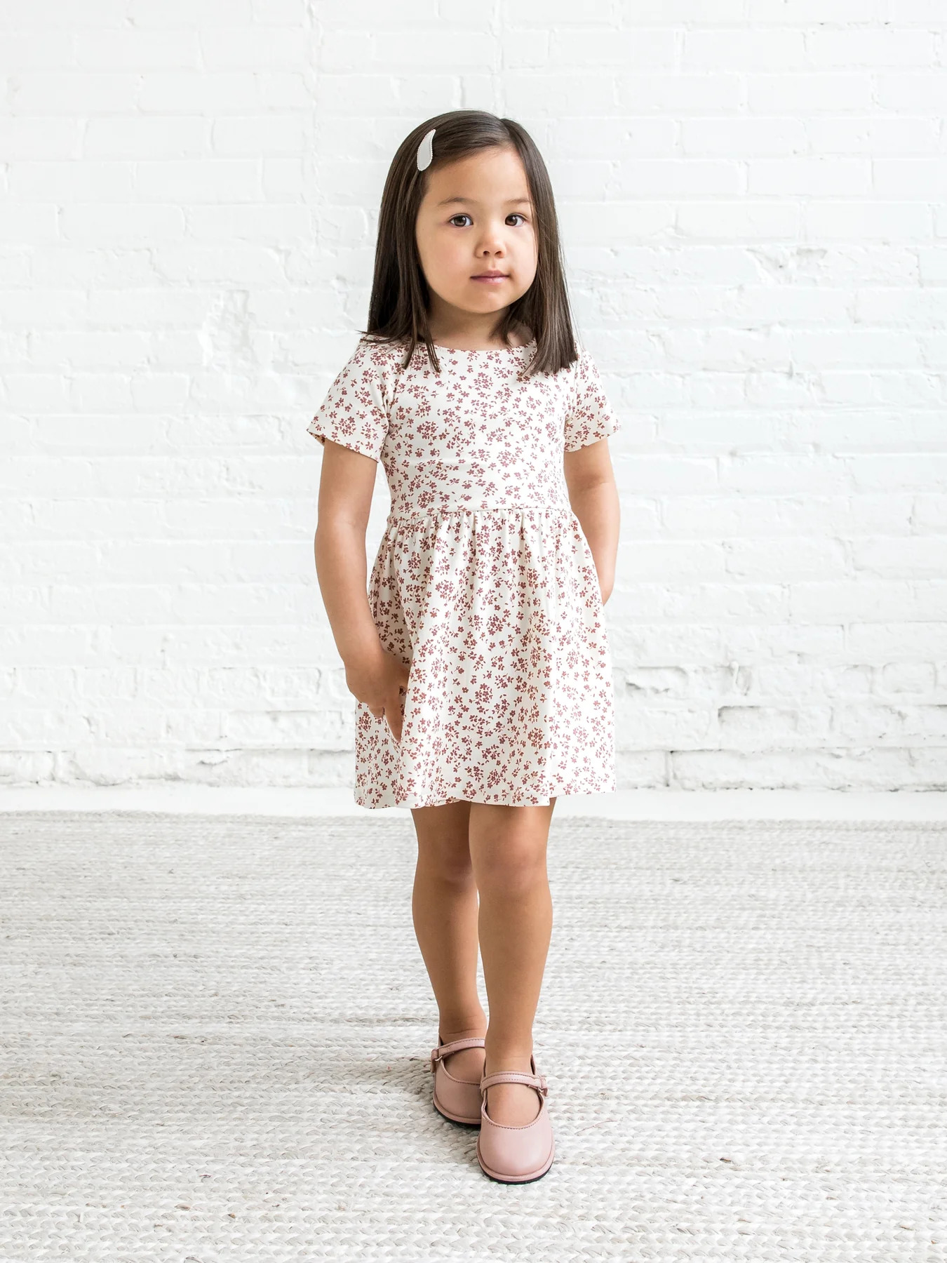 A little girl in a pink floral dress standing in front of a white wall.
