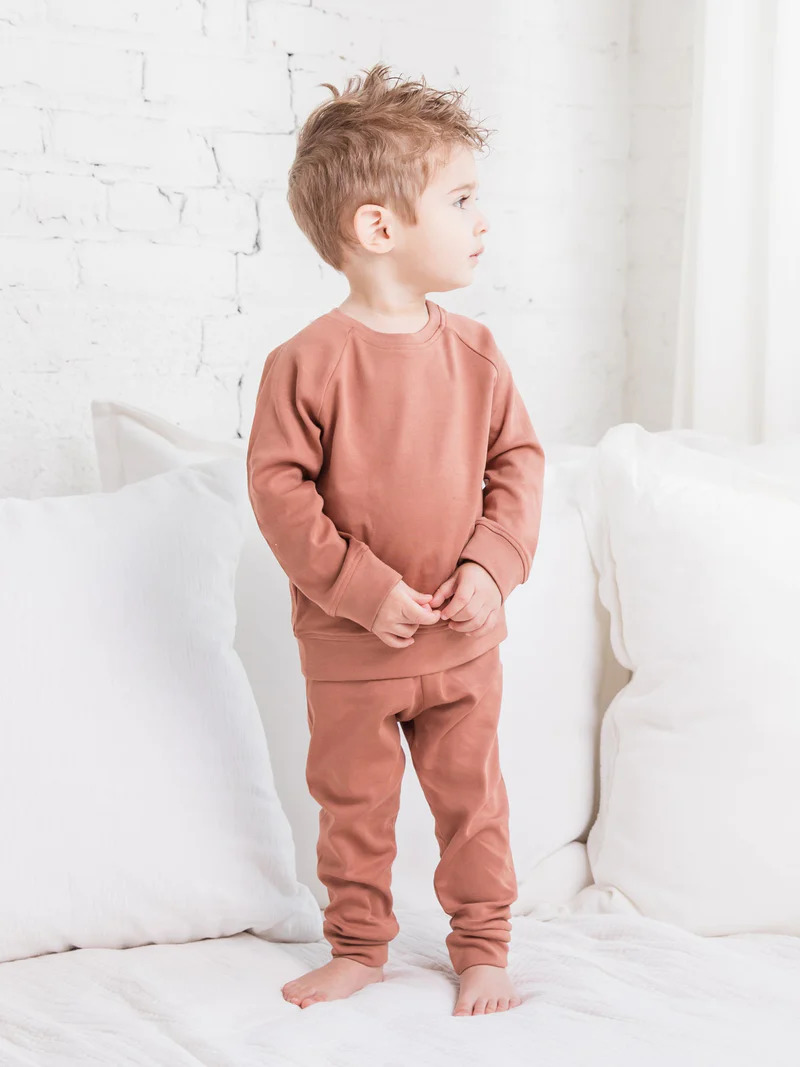 A young boy standing on a bed wearing a brown sweatshirt and pants.