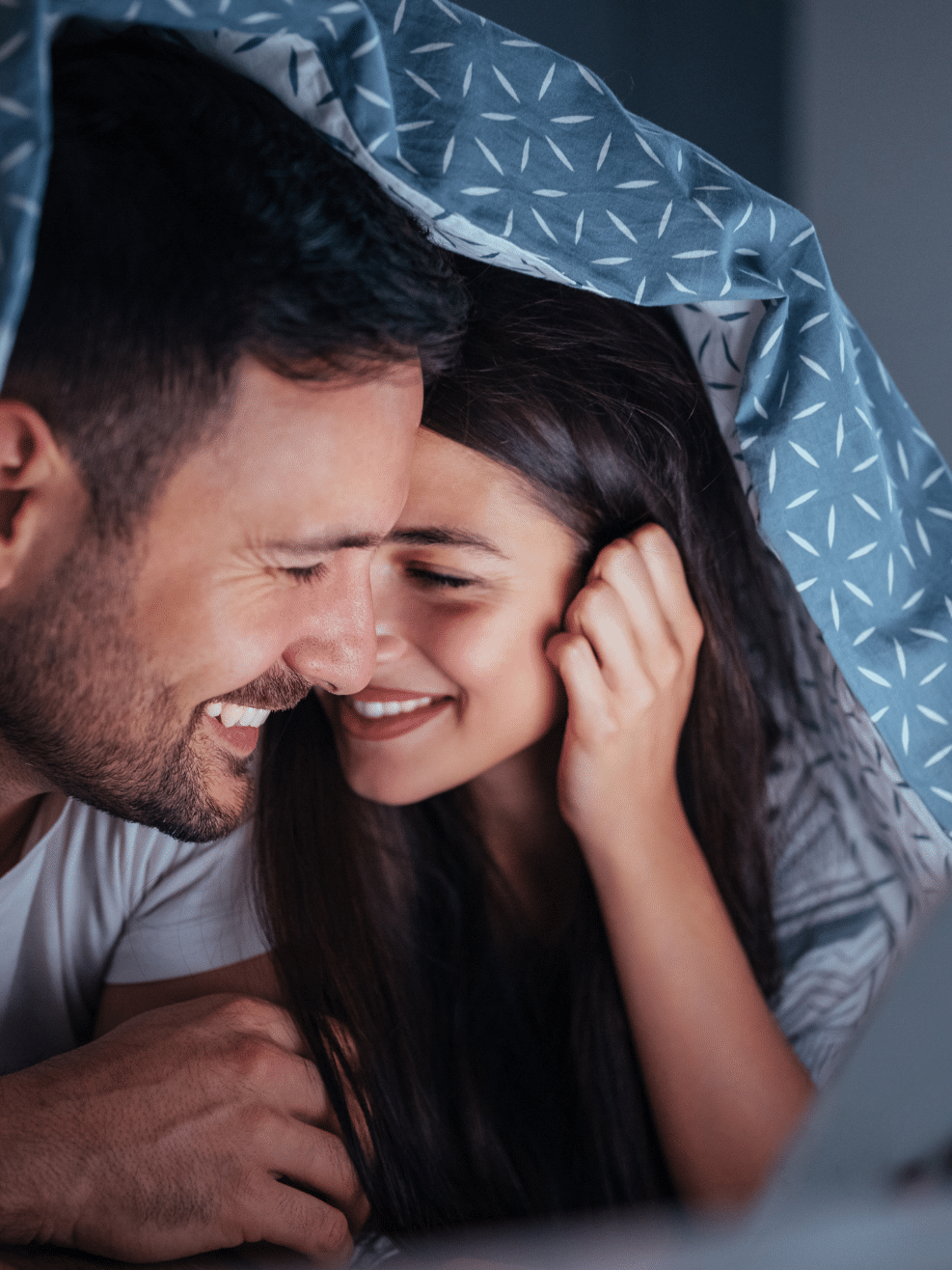 A man and woman are lying under a blanket and using a laptop.