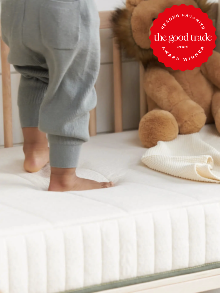 Child standing on a mattress in a crib beside a teddy bear and blanket. Red badge in corner says "the good trade 2025 award winner.