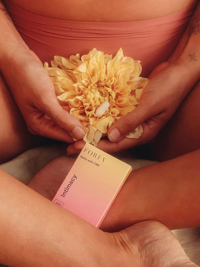 Close up of a model seated with their legs crossed, holding a flower, with Foria's Intimacy Suppositories under their hands.