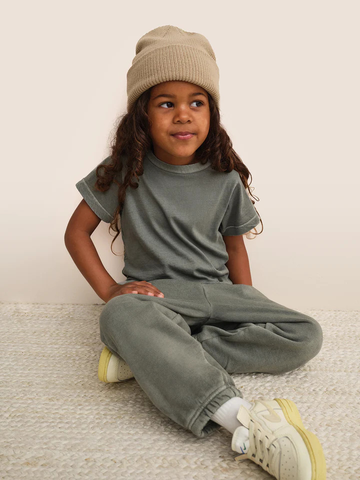 A little girl sitting on the floor wearing a beanie and sweatpants.
