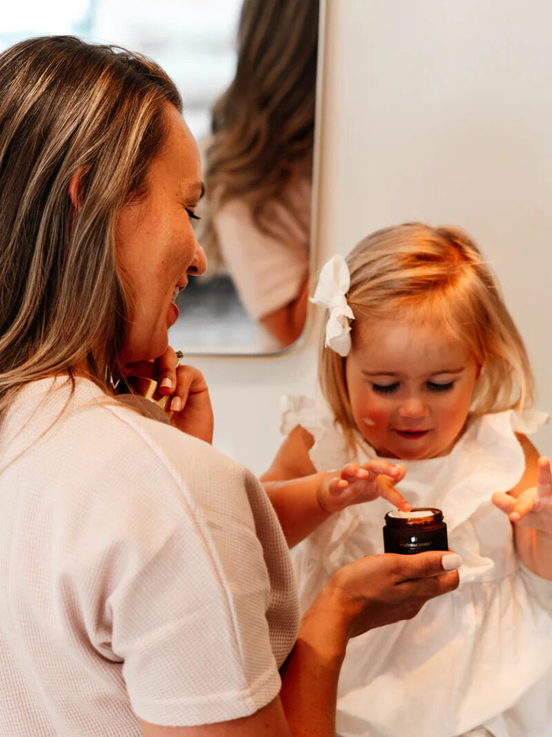 A model holding a jar of Matrescence cream while a child dips their finger in it. 