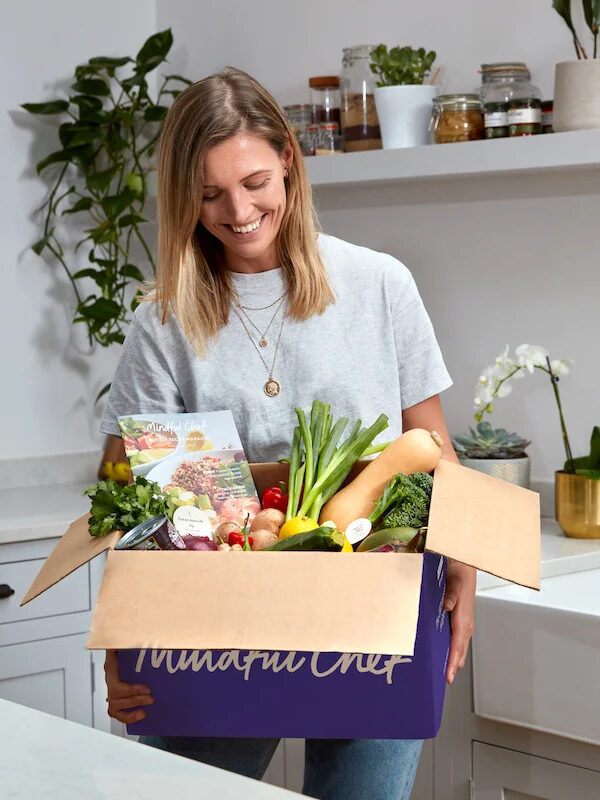 A model in a kitchen holding an open Mindful Chef meal delivery box. 