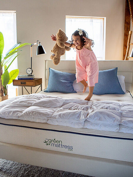 A child jumping on a My Green Mattress while reaching for a teddy bear, mid-air. 