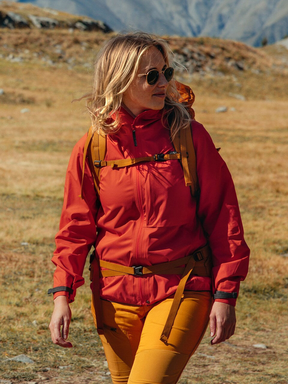 A woman in a red jacket walking through a field with mountains in the background.