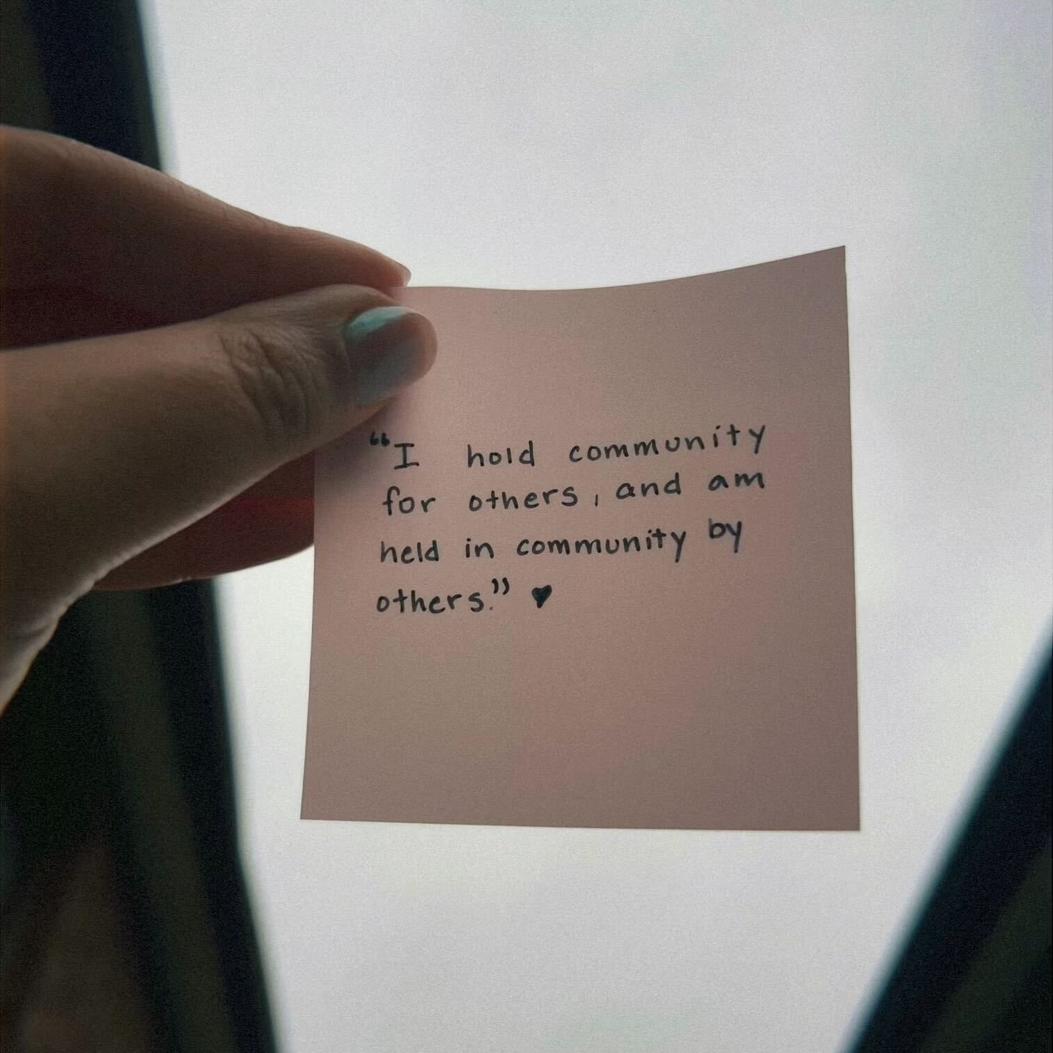 A sticky note being held up to the sky, reading "I hold community for others, and am held in community by others".