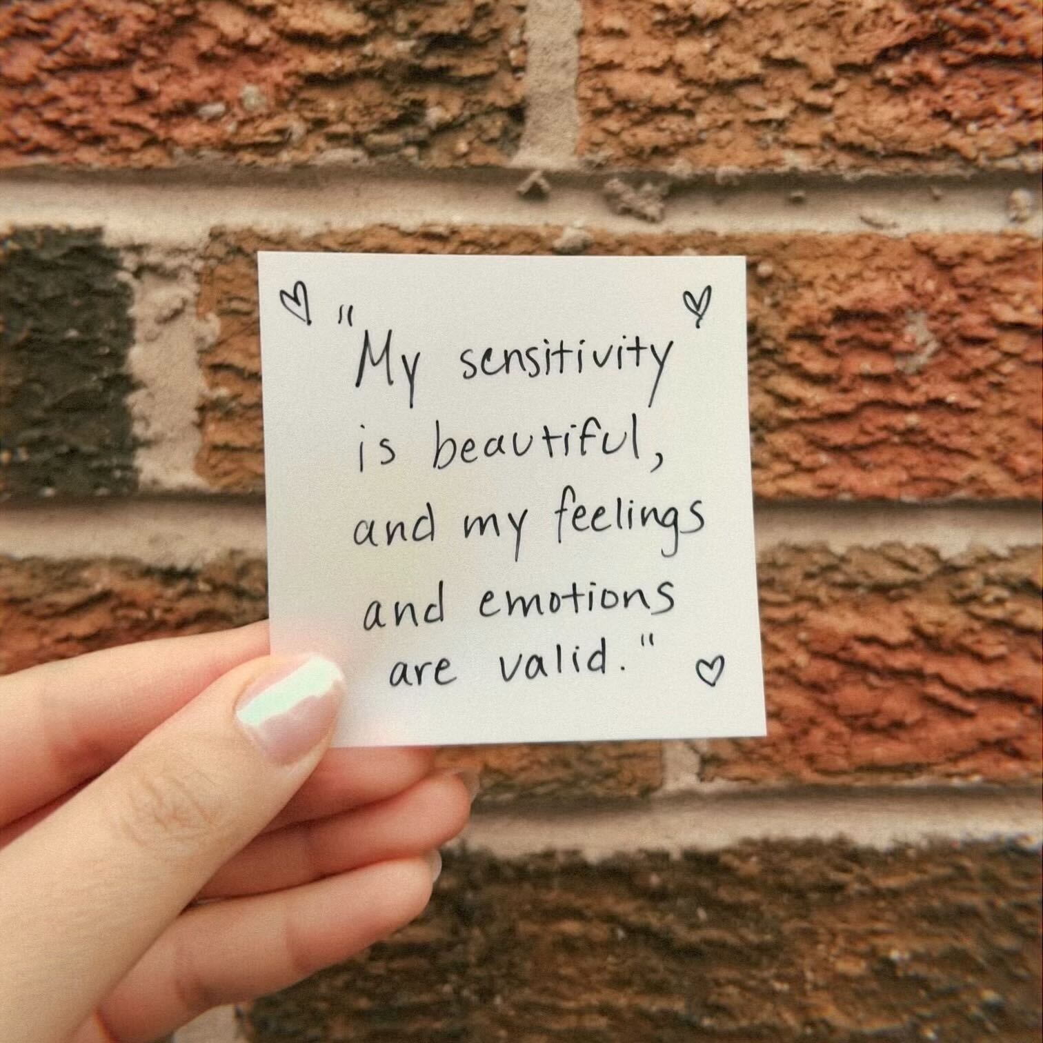 A sticky note held up in front of a brick wall, reading "My sensitivity is beautiful, and my feelings and emotions are valid". 