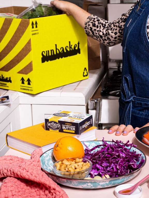 A close up of a model in a kitchen reaching inside a Sunbasket delivery box, with a plate of foods from the box set in front of them on a kitchen table.