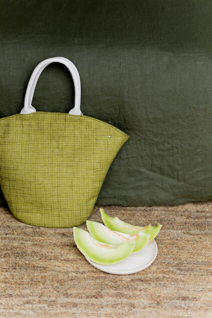 Green woven tote bag with white handles on a bench and a plate with sliced honeydew melon beside it.