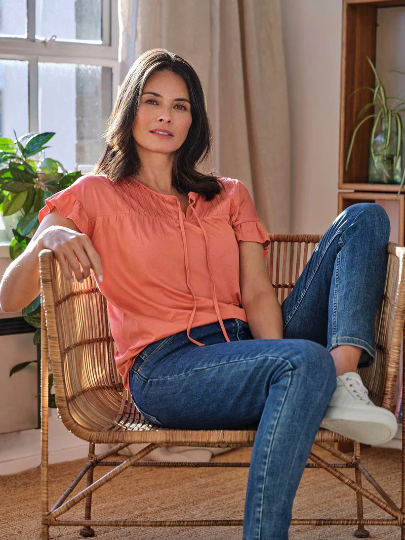 A woman sitting in a wicker chair wearing a pink top and jeans.