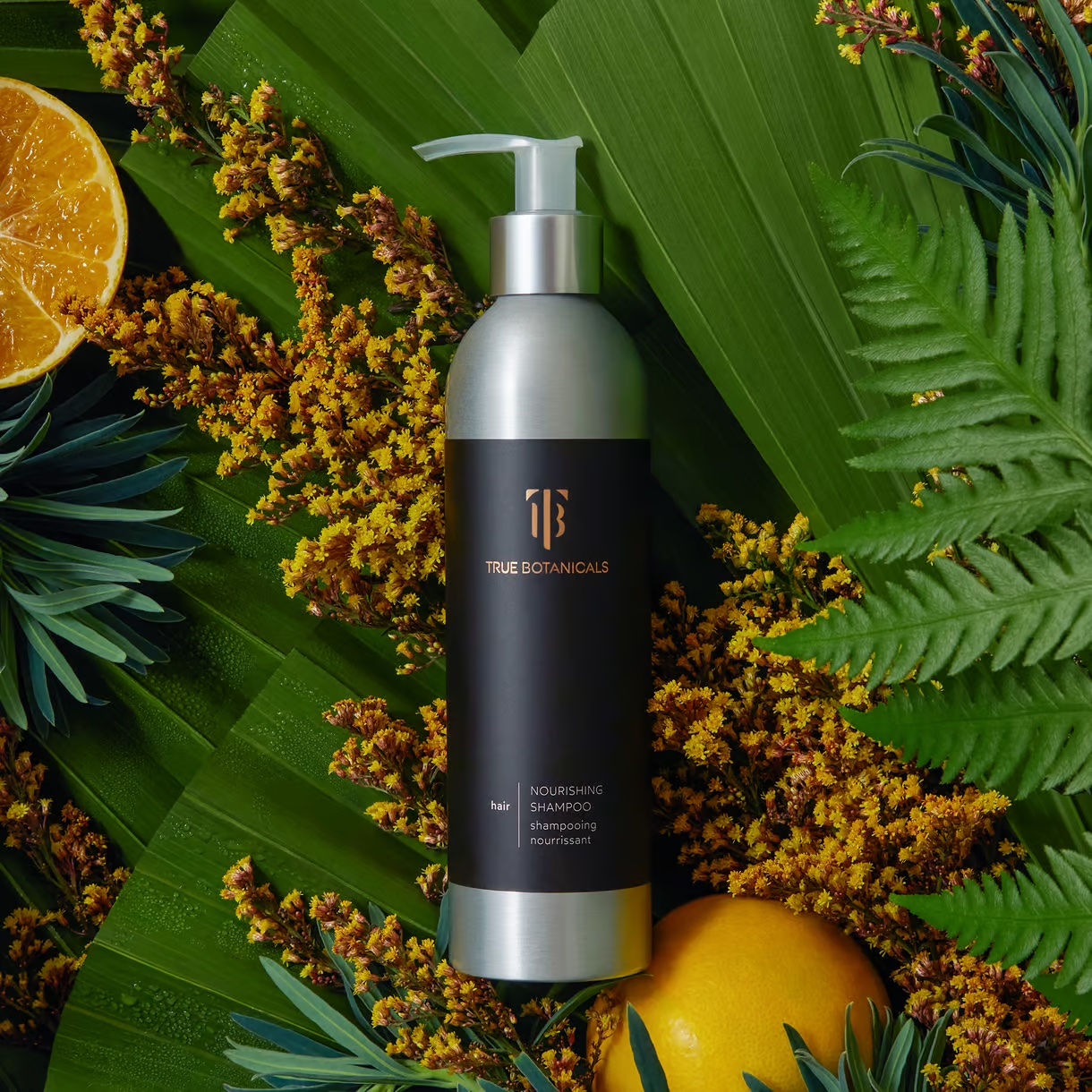 A bottle of true botanicals nourishing shampoo surrounded by citrus slices and yellow flowers against a backdrop of green leaves.