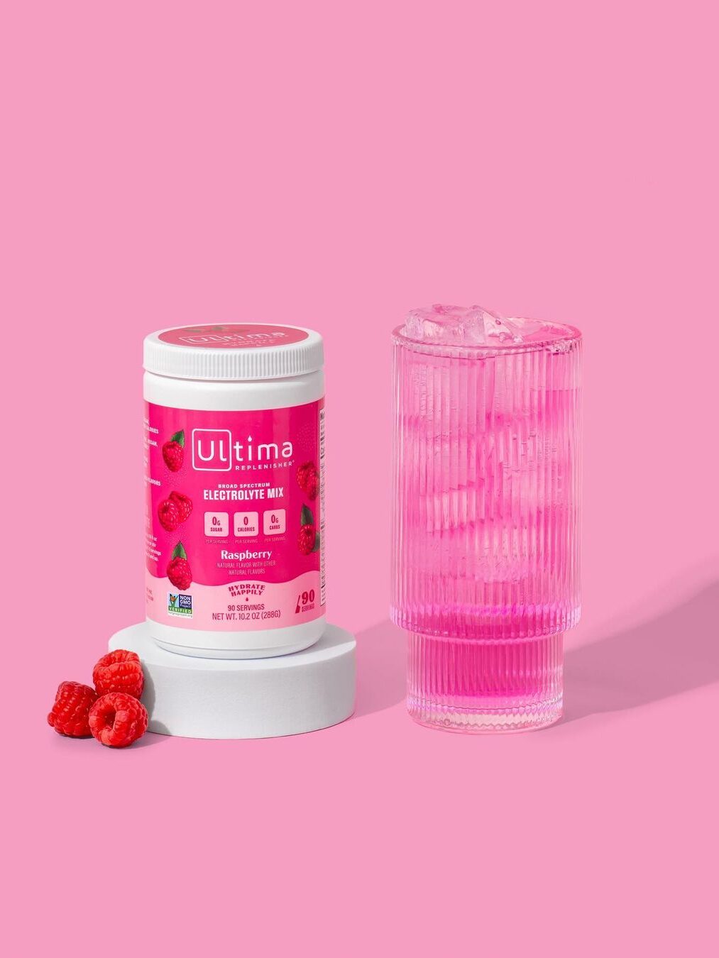A container of Ultima Raspberry Electrolyte powder on a stand next to a tall glass of the ready-made electrolye drink.