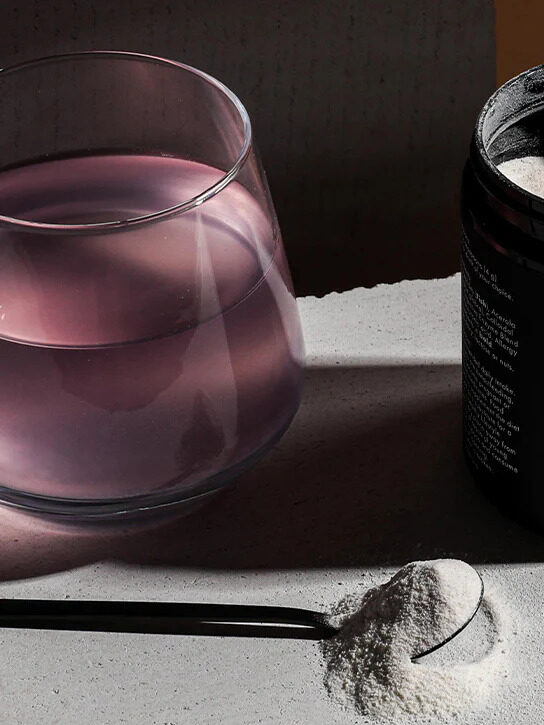 A glass filled with a clear purple drink, with an open jar of WelleCo collagen next to it and a spoon of collagen powder in front. 