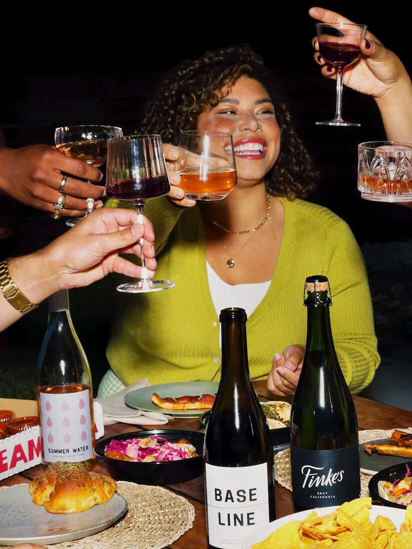 A group cheers with wine glasses at a dinner table, with a model in the center. Wines are from Winc. 