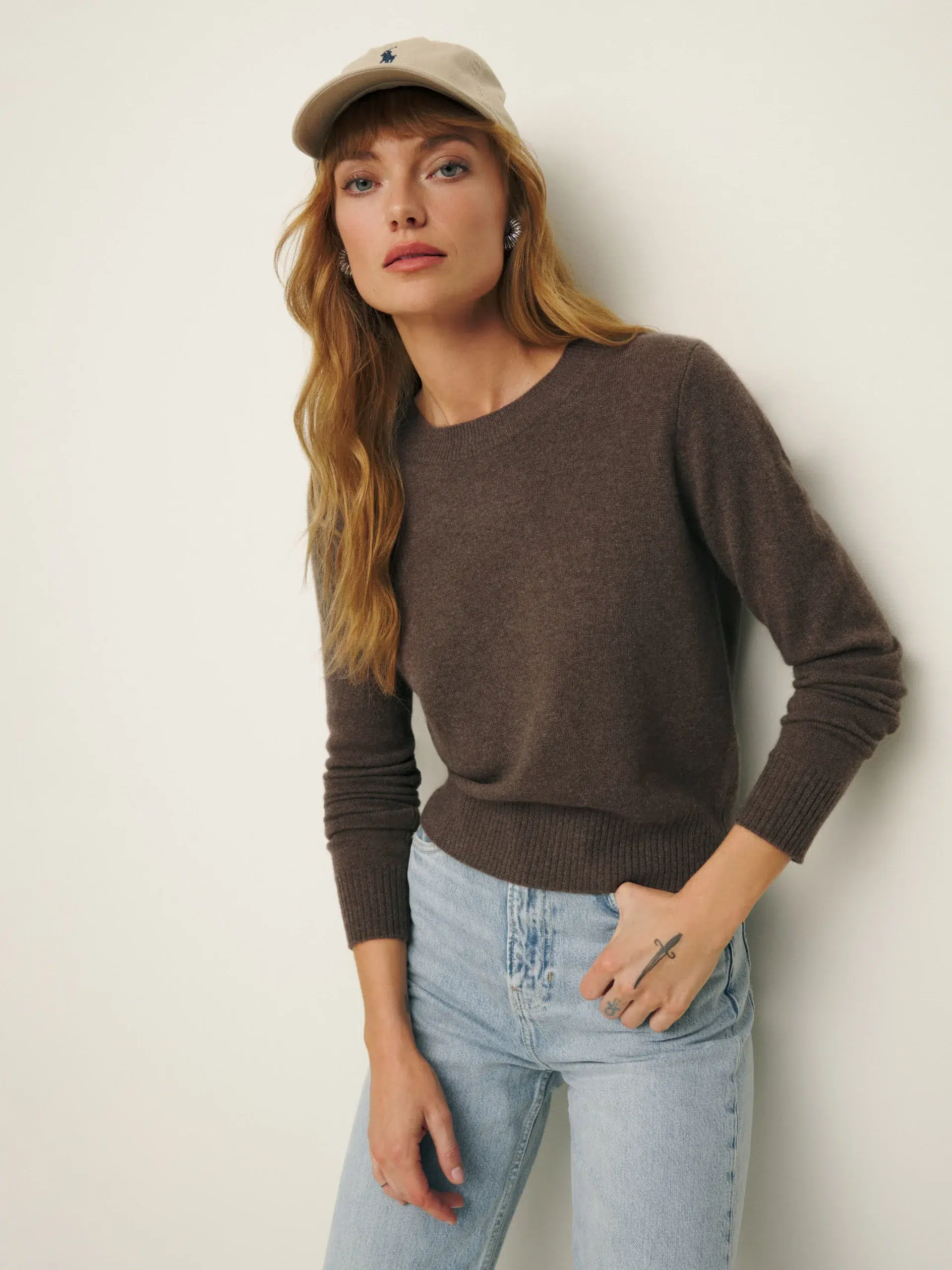 Woman in a brown sweater and blue jeans posing with her hand on her hip and wearing a beige cap.