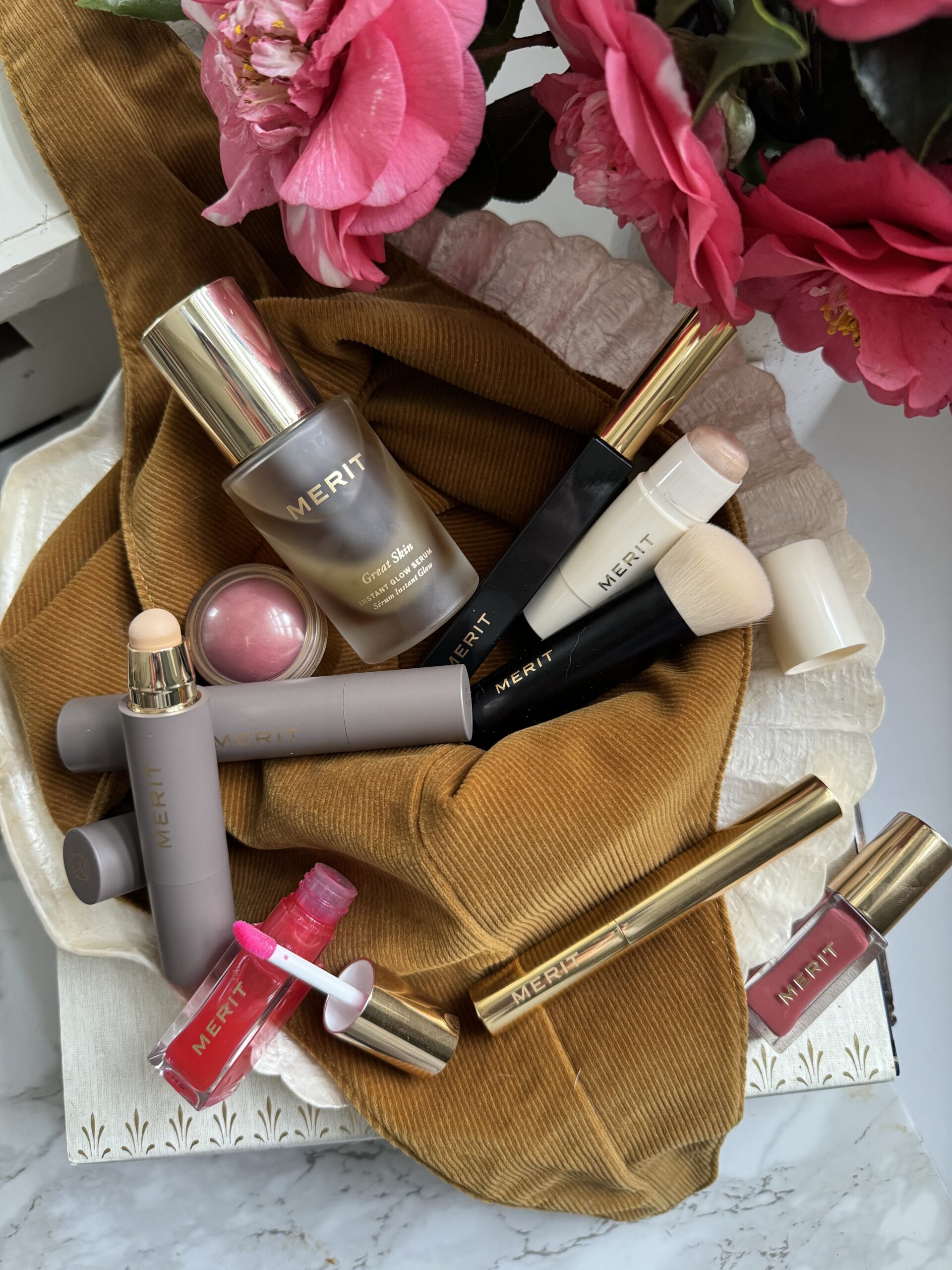 Assorted cosmetic products arranged on a draped earth-toned fabric with pink flowers nearby.