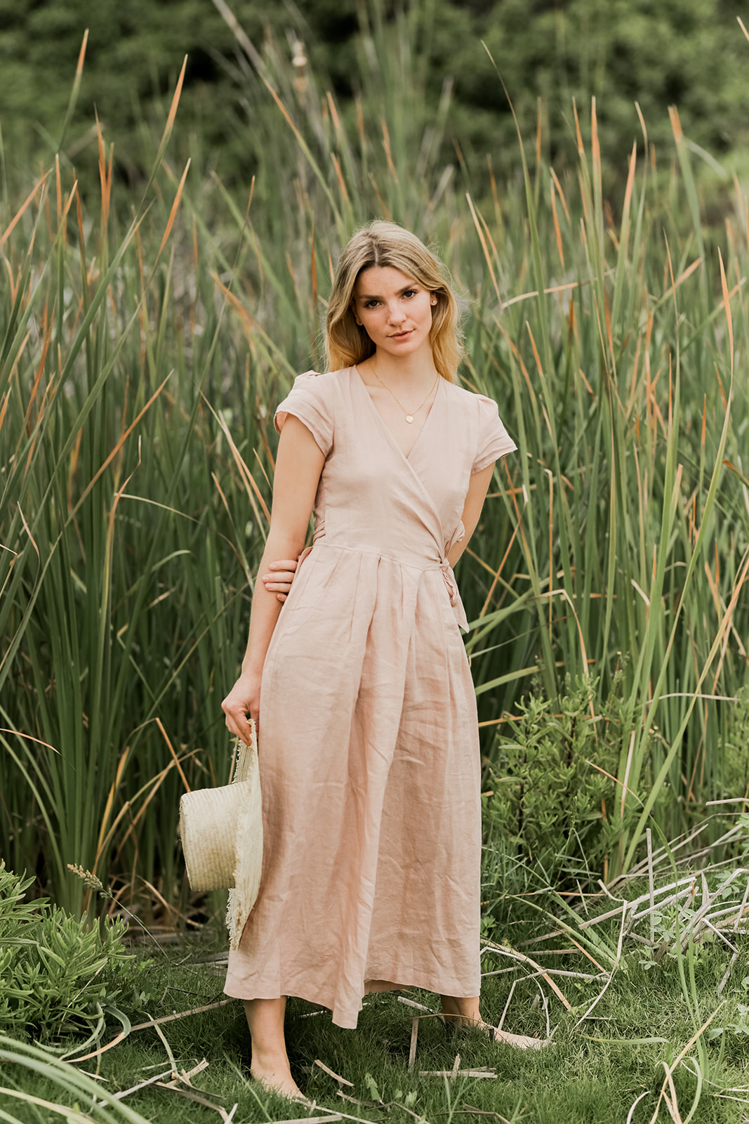 A woman in a beige dress standing among tall grasses with a straw bag at her side.