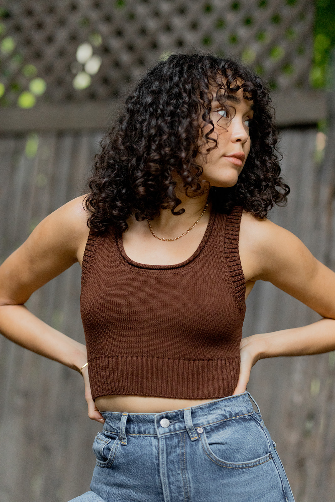 A woman with curly hair wearing a brown tank top and blue jeans standing with hands on hips.