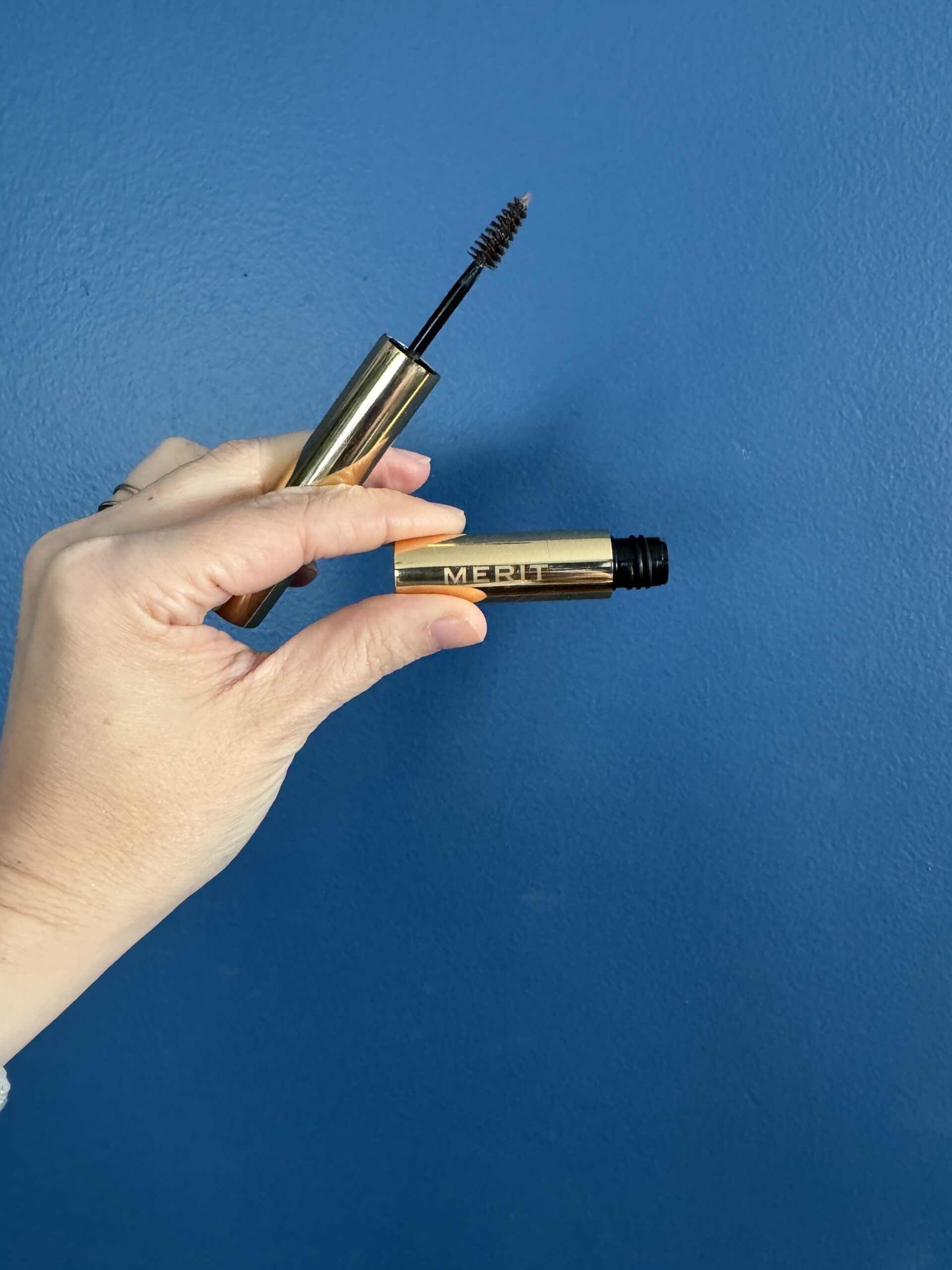 A hand holding an open mascara tube against a blue background.